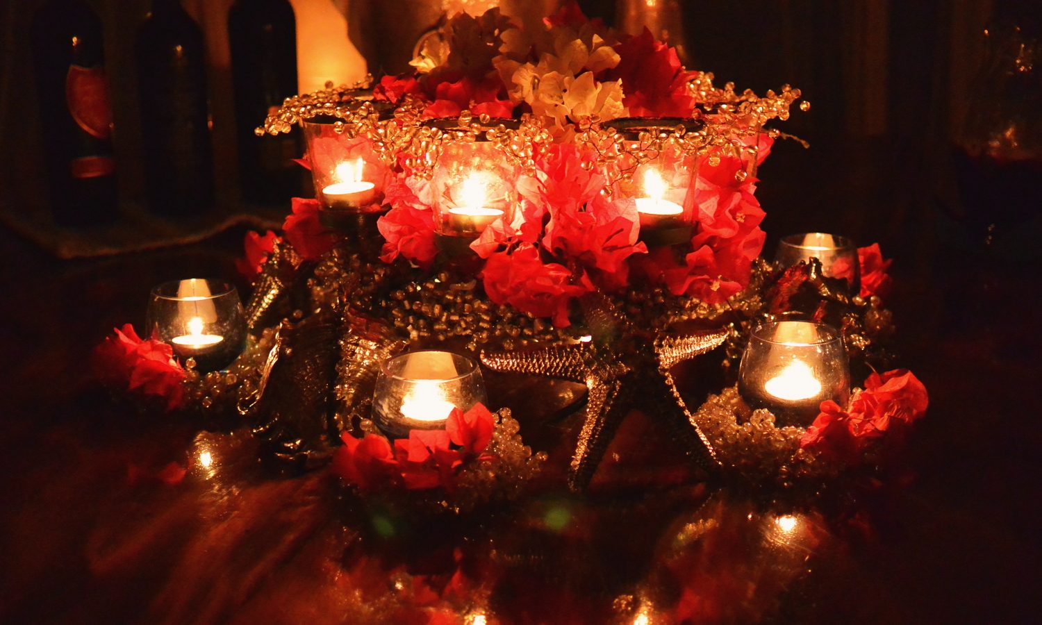 A dining table centrepiece of red flowers and votive candles glowing in the dark