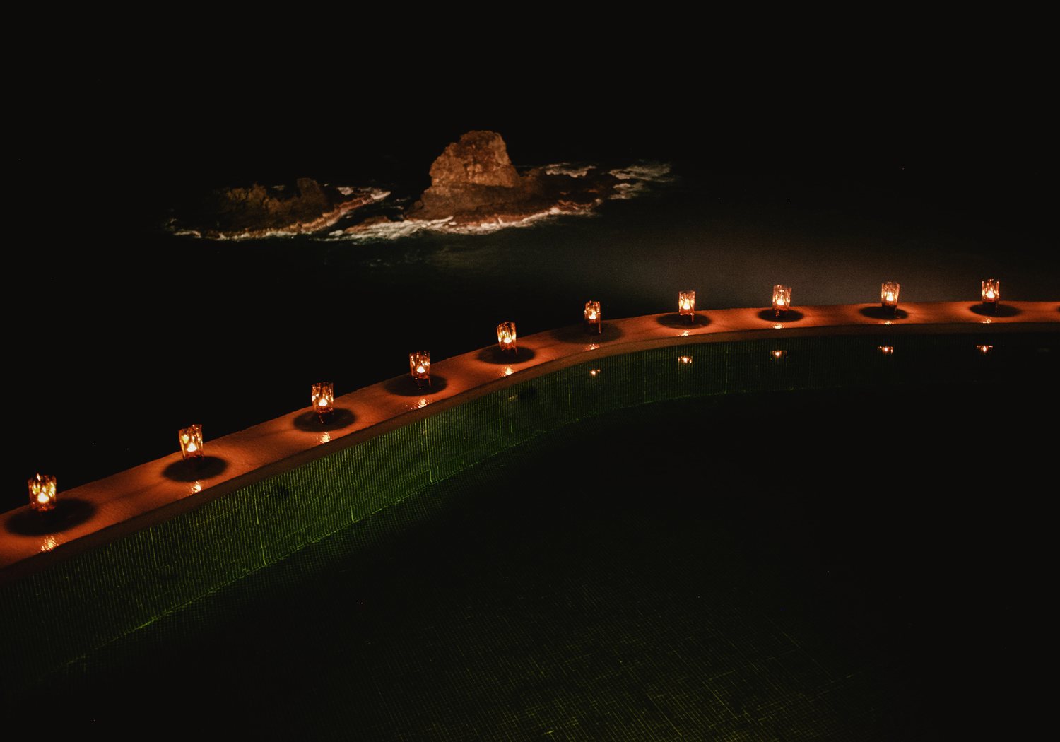 Candles in glass holders along the curved edge of the pool at night, with a lit sea rock and surf beyond