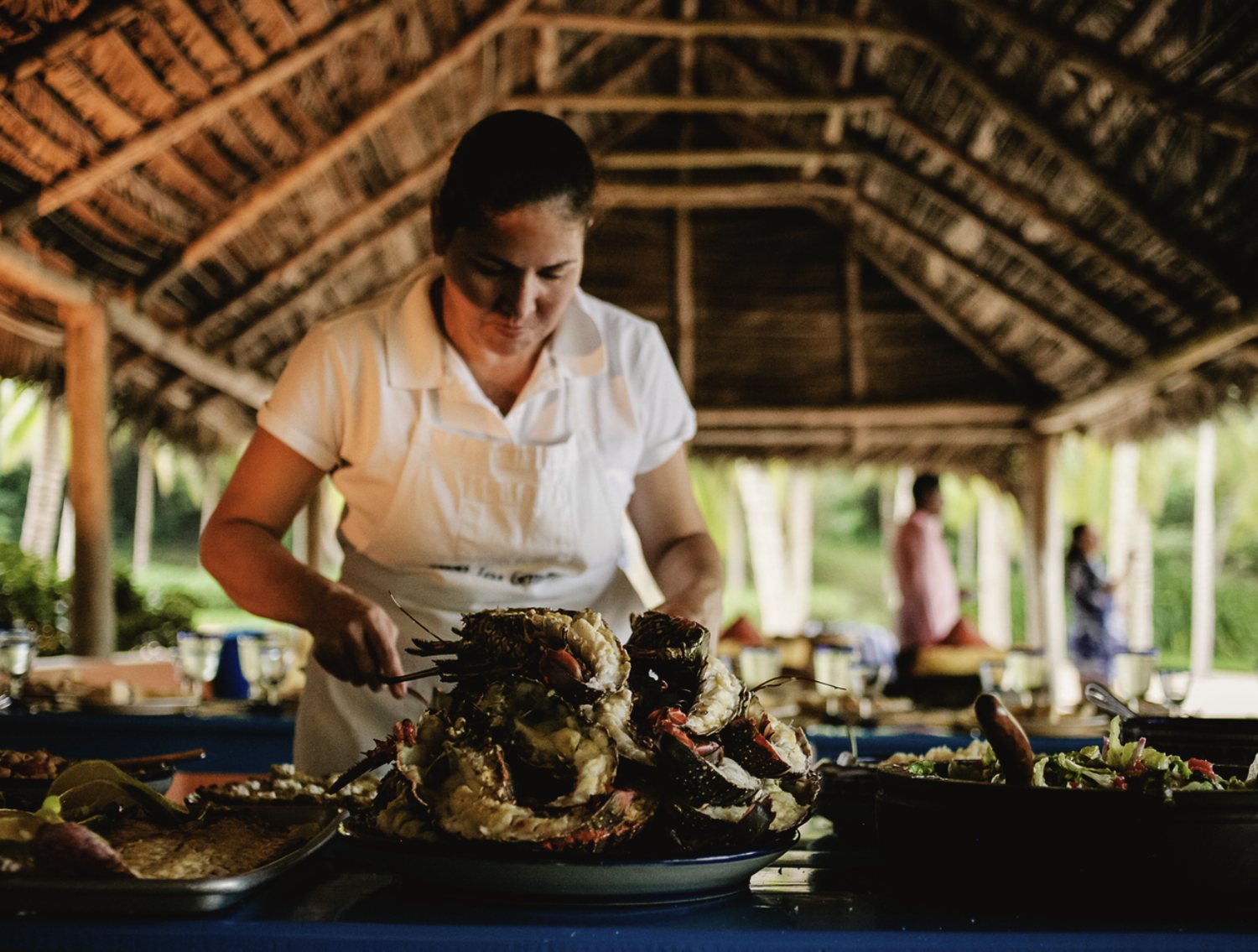 The chef in a white apron plating seafood at a long table under the thatched palapa