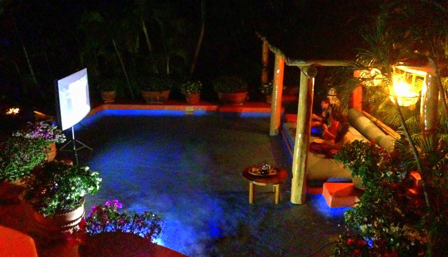 Outdoor movie screen beside a glowing blue pool, with a palapa lounge area and potted flowers at night