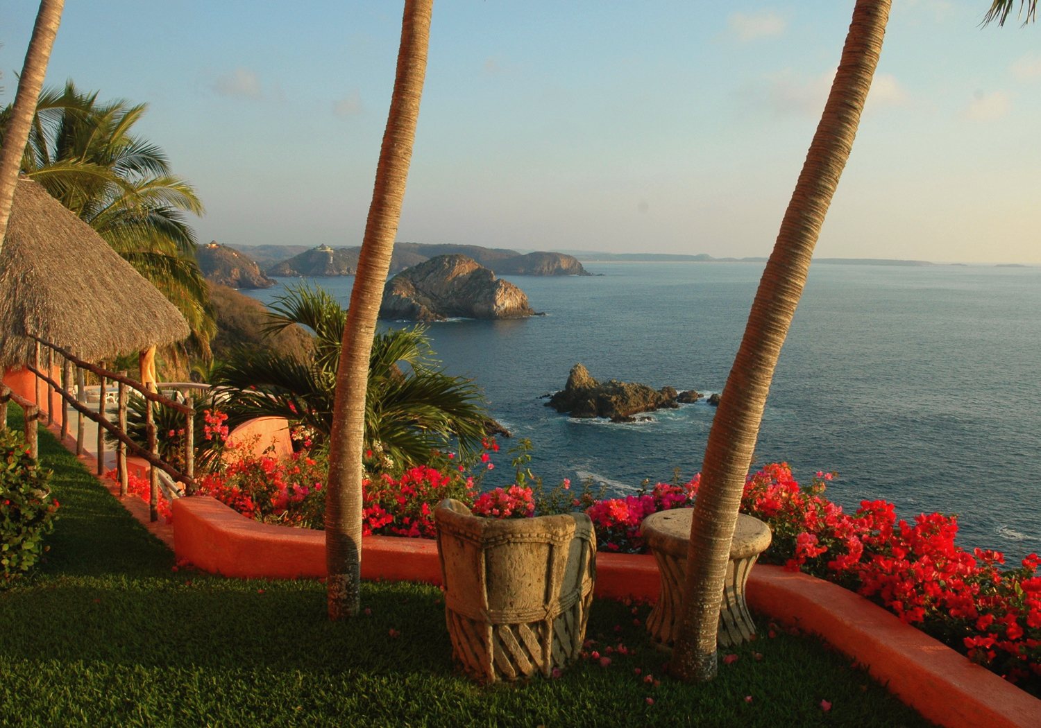 Palms and red bougainvillea on the cliff lawn, with a thatched roof, the bay and sea stacks beyond