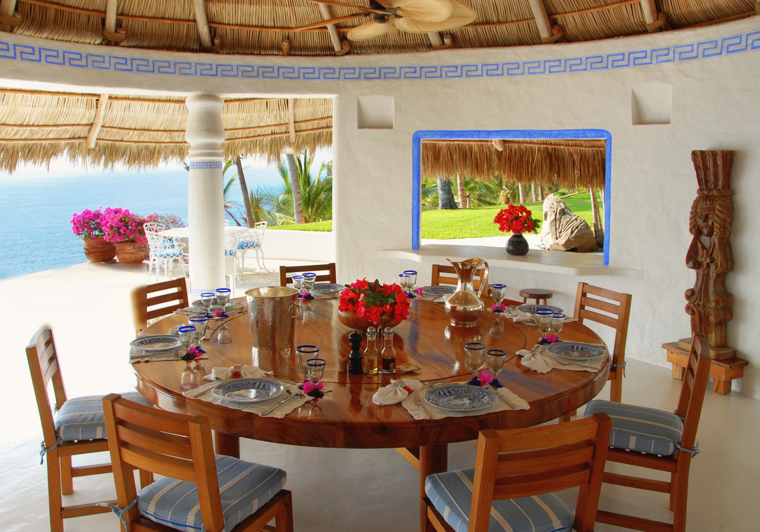 The palapa dining room by day: a round wooden table set for a meal beneath a thatched roof, with the sea and garden visible through the openings