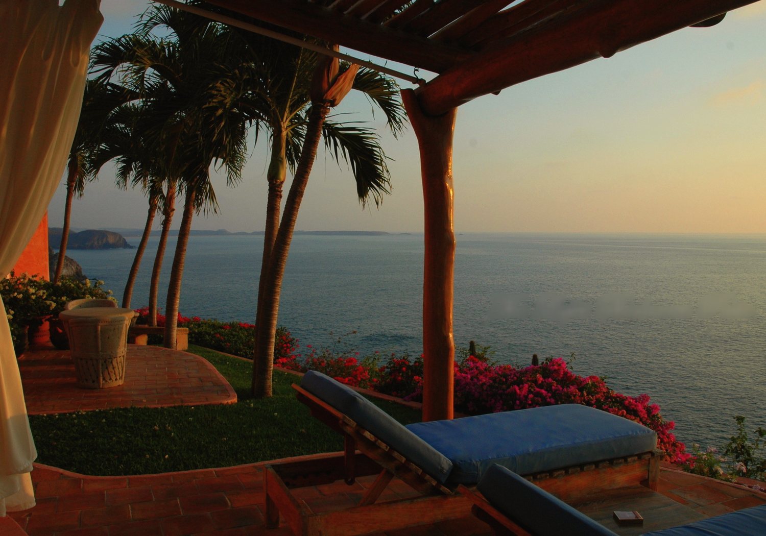 The Lapis patio at sunset: a pergola, palms and a blue-cushioned lounger above the ocean