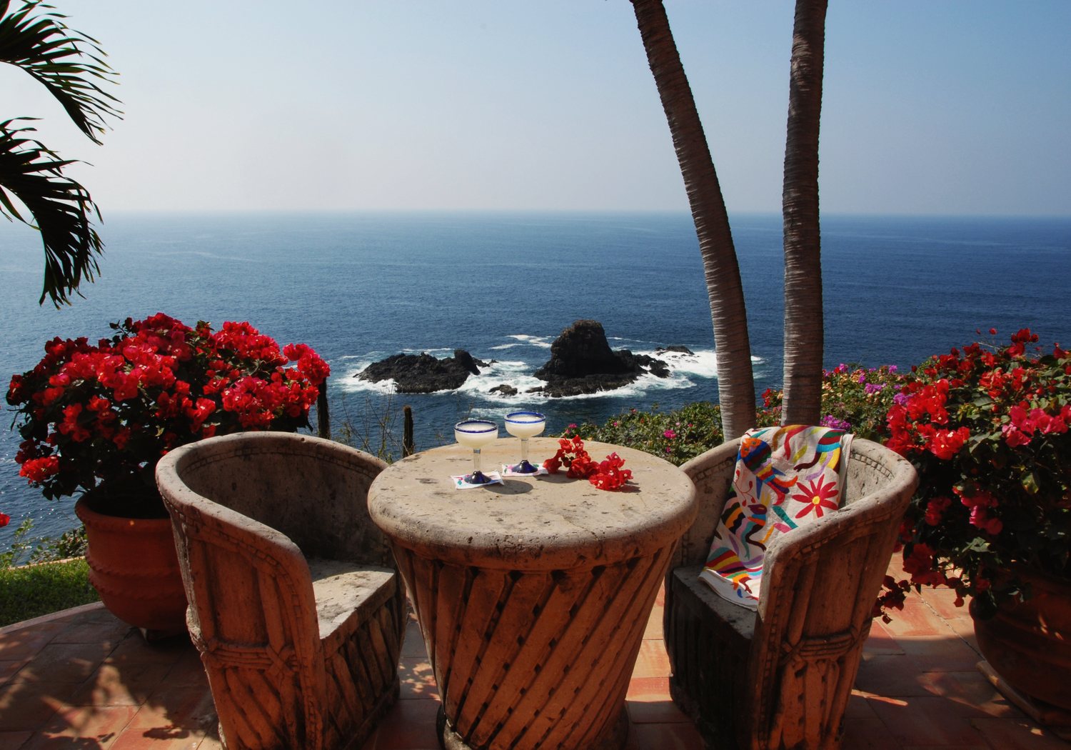 The view from the Lapis terrace: a stone table and carved chairs among red bougainvillea above the ocean and rocks