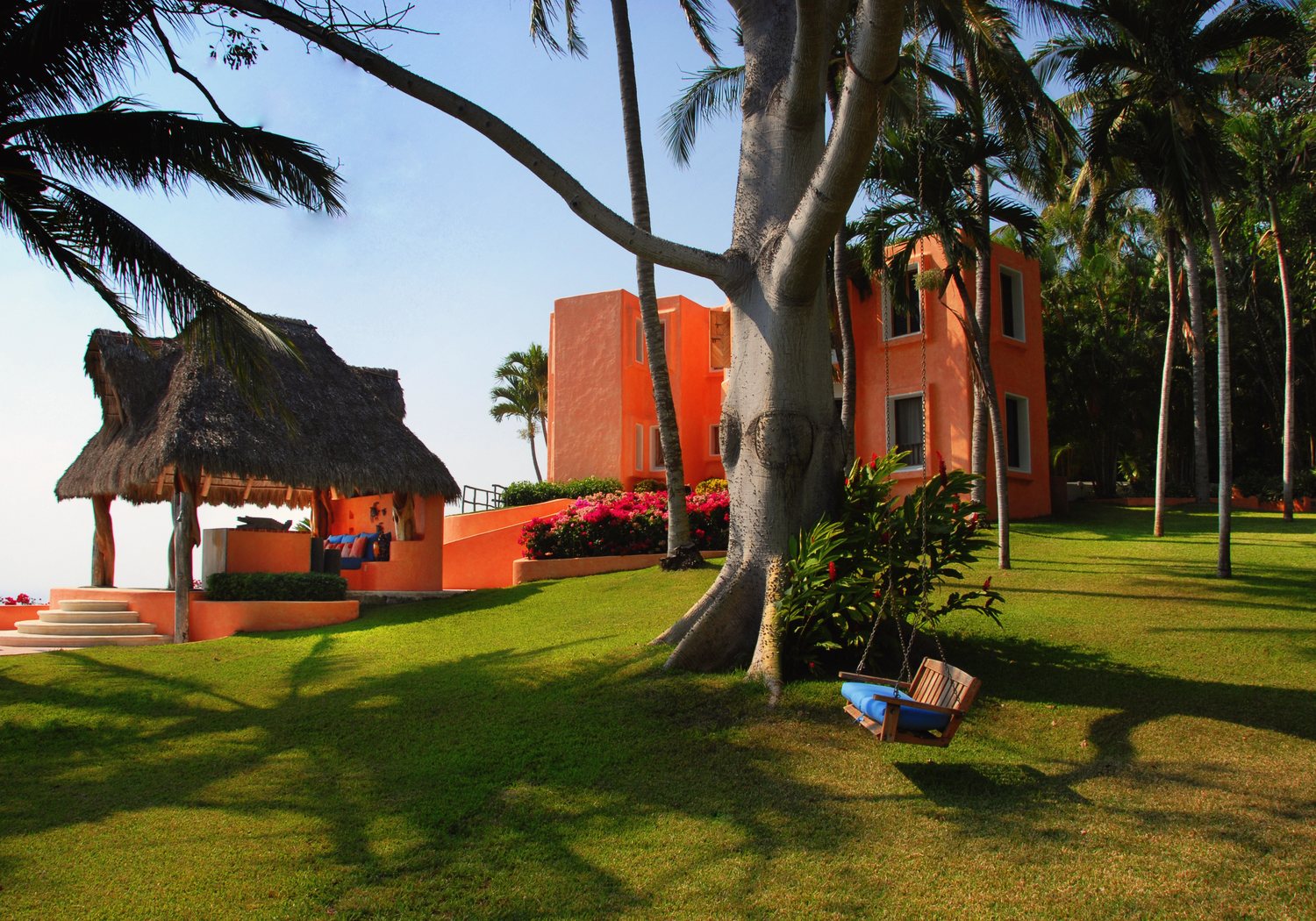The lawn: the thatched massage palapa on the left and the orange Emerald room building beside a large ceiba tree with a swing, palms and bougainvillea