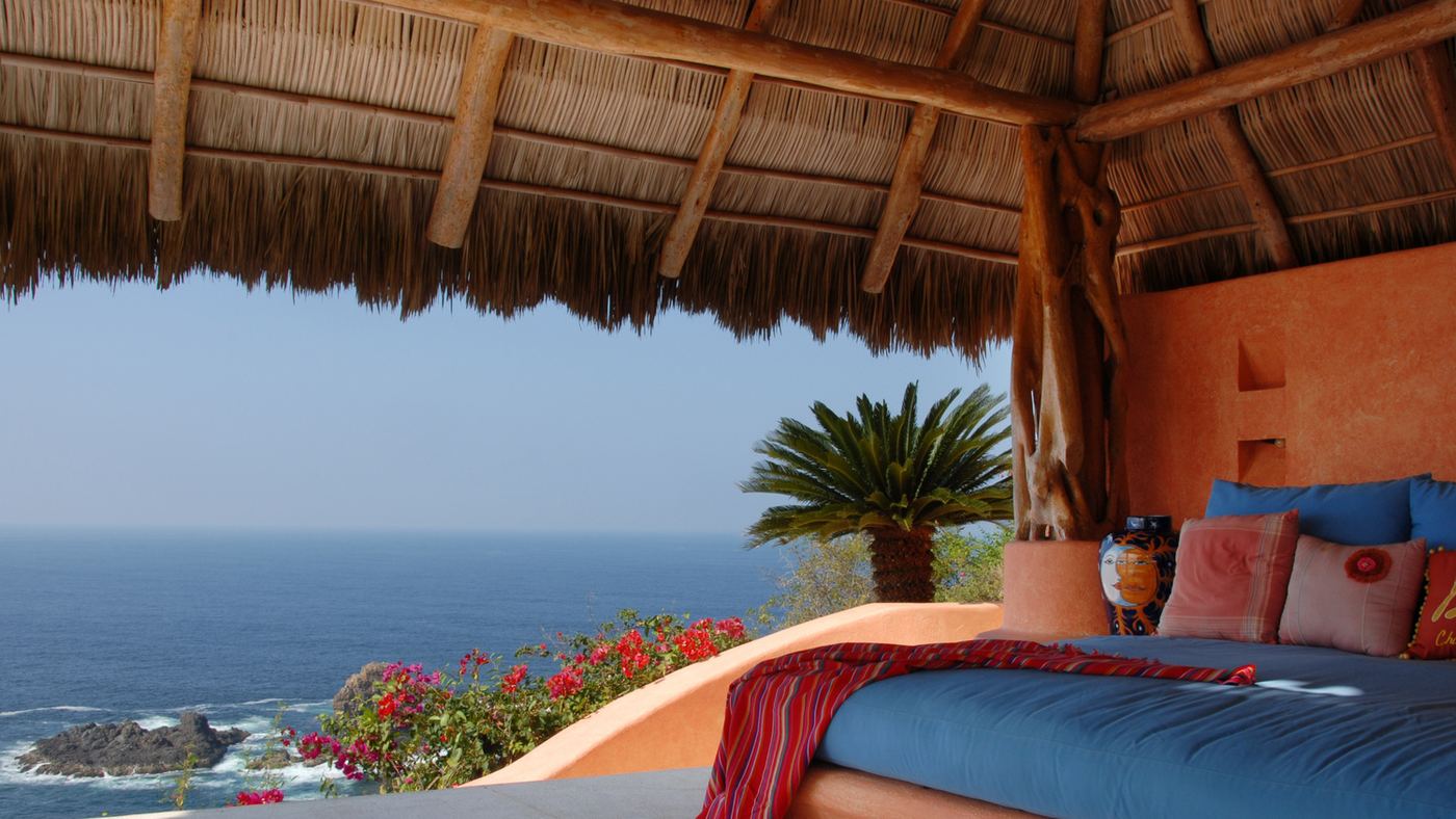 The open-air massage palapa: a blue daybed under a thatched roof with terracotta walls, looking out to the Pacific