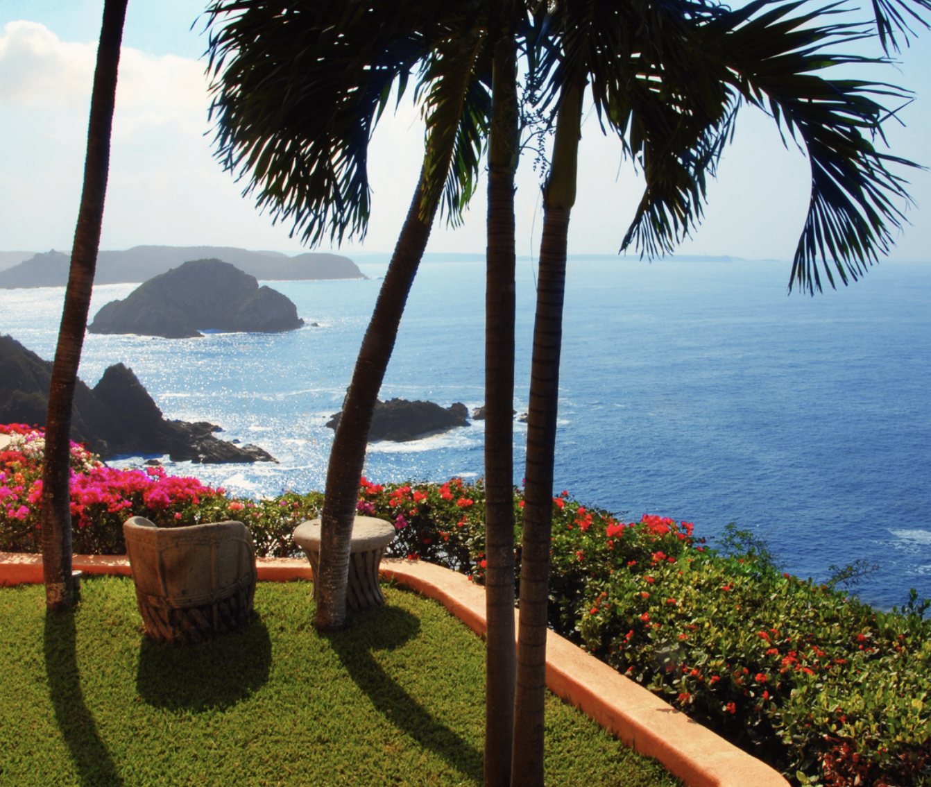 The lawn outside Pariba: a carved seat and stone table among palms and bougainvillea, above the sea and rocks