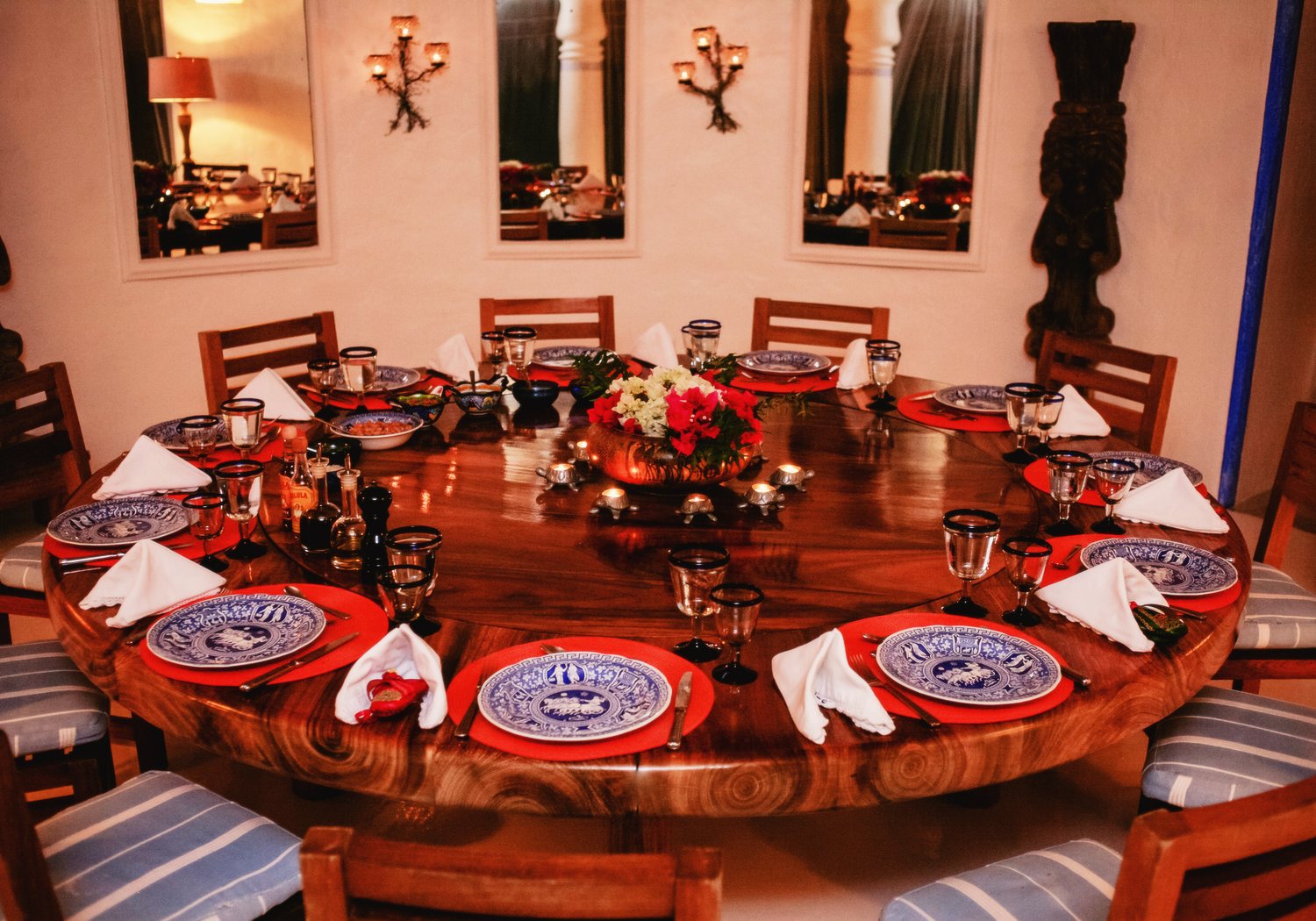 A round wooden table in the palapa set for dinner at night, with blue-and-white plates on red placemats, a centrepiece of flowers and small candles, and mirrors on the wall behind