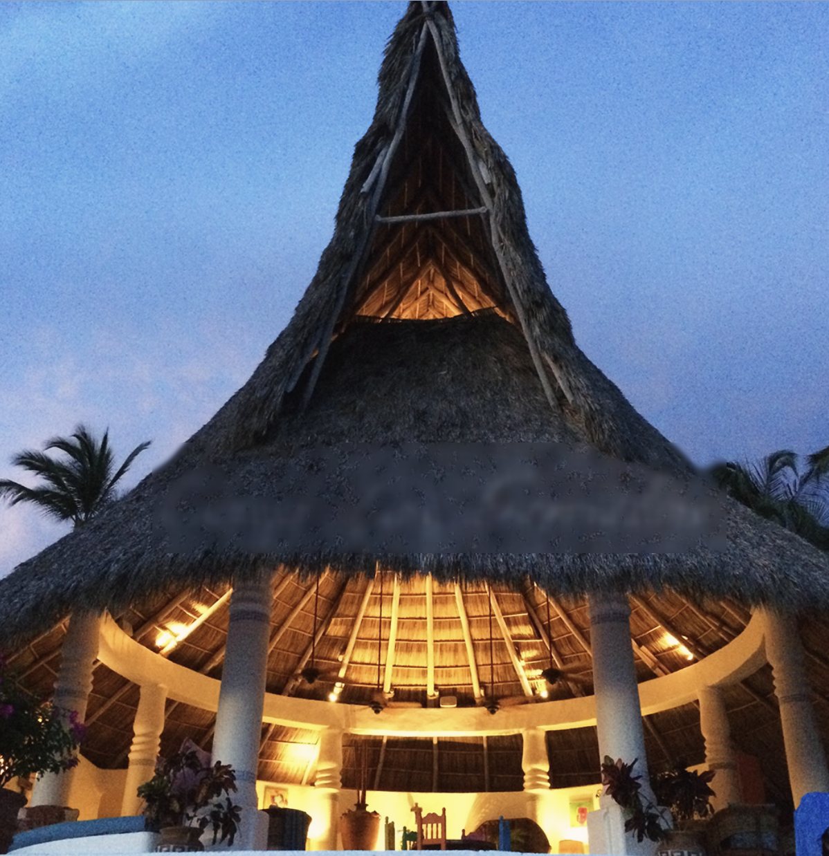 The tall thatched palapa lit from within at dusk, seen from the pool