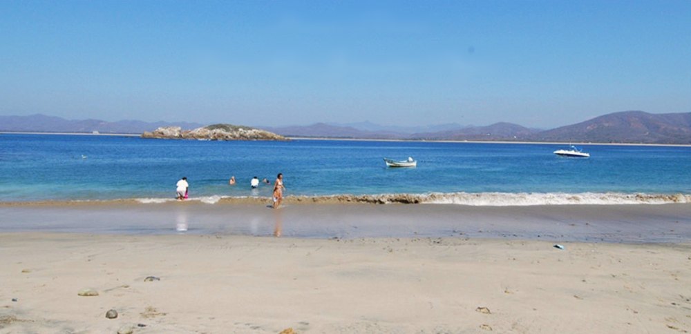 A wide sandy beach beside a blue bay, with swimmers at the water's edge, boats anchored and an island and hills beyond