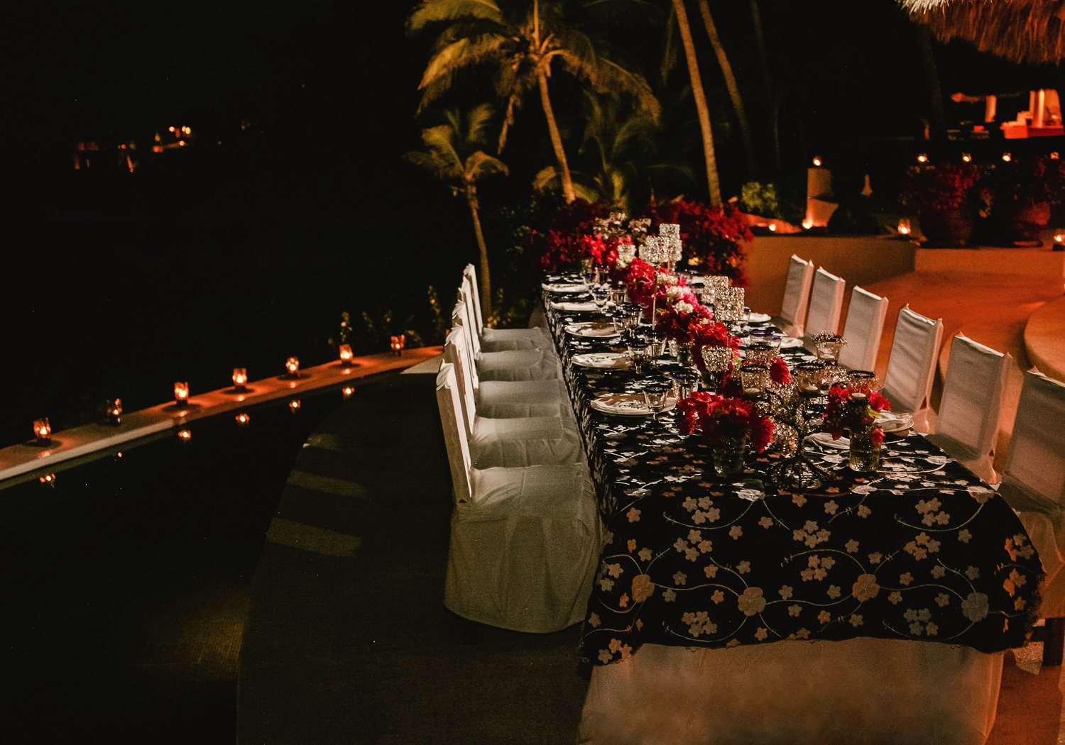 A long table dressed in black floral linen with red flowers beside the pool at night, candles along the pool edge