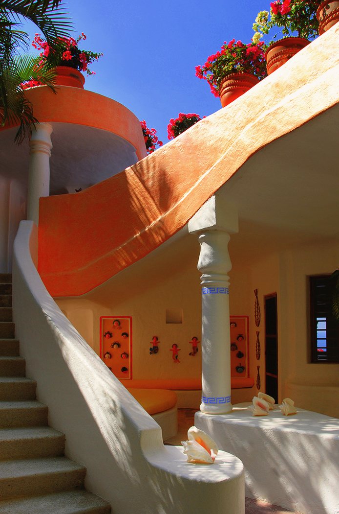 The white staircase beside an orange curved wall, with flowering pots above and shells on a low wall