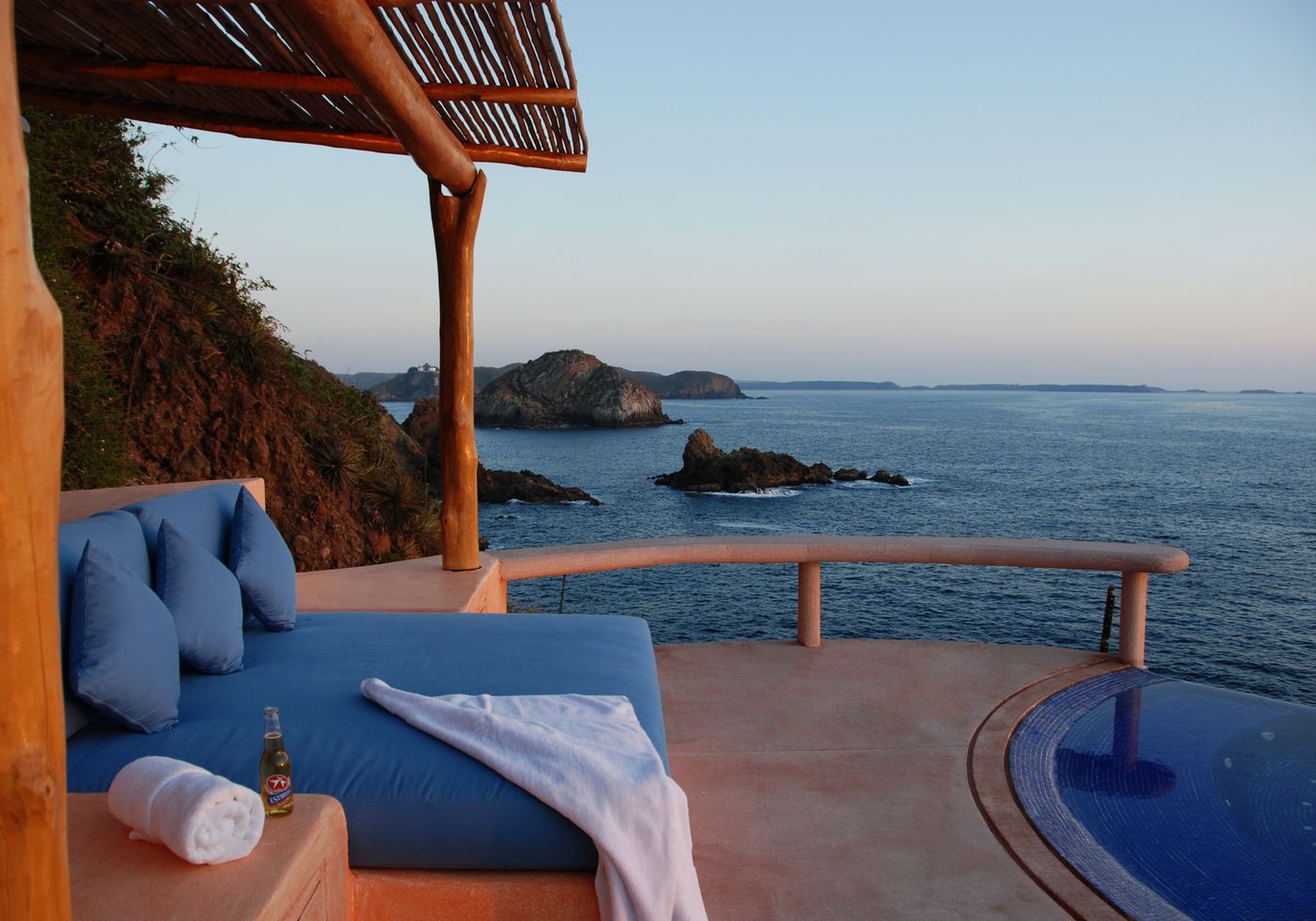 A daybed with blue cushions and a rolled towel under a thatched pergola at sunset, with the ocean and rocky islets beyond