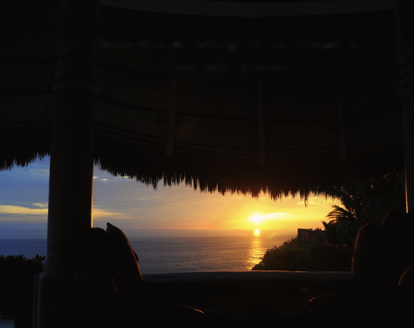 The sun low over the sea, seen from beneath the thatched roof of the living room palapa between white columns